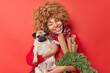 Happy curly haired woman has friendly relationships with favorite pug dog holds green wreath and rolled paper wears sweater and scarf around neck isolated over red background. New Year time.