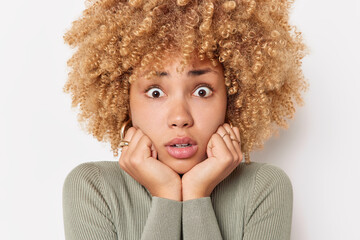 Wall Mural - Scared woman with curly hair keeps hands under chin stares with worried expression cannot believe her eyes wears casual jumper isolated over white background. Surprised excited female model.