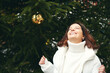 © annanahabed - Outdoor portrait of happy woman next to green Christmas tree, wearing white pullover and jacket