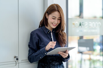 Wall Mural - Front view happy successful young Asian woman holding a pen using a tablet standing at the office.