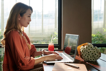 Wall Mural - Smiling young businesswoman working on laptop in cafe, answering e-mails and filling documents