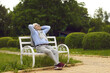 © Studio Romantic - Happy senior man relaxing in a green summer park. Retired chubby old man smiling, breathing in fresh air and looking at the sky while sitting with hands behind head on wooden bench in a city garden