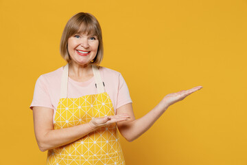 Elderly happy fun housekeeper housewife woman 50s in orange apron point index finger aside on workspace area isolated plain on yellow background studio portrait. People household lifestyle concept