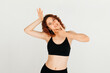 © ChesterAlive91 - Cheerful woman with curly red hair smiling, posing with her hand near her face in black sport top, standing at white background
