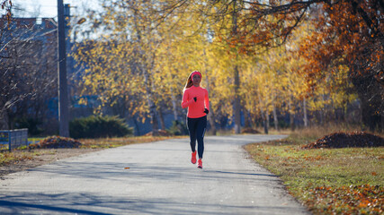 Wall Mural - Young fitness woman running on the road in the cold morning in winter. Sporty girl in orange long-sleeve jogging in the sunny morning