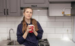 © Наталья Анюхина - A young brunette girl stands in the kitchen alone in the morning for breakfast and drinks from a red cup with a smile on her face