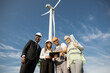 © sofiko14 - Group of four multiracial coworkers in safety helmets smiling on camera while staning on farm with huge windmills. Men and women using digital tablet and blueprints during meeting outdoors.