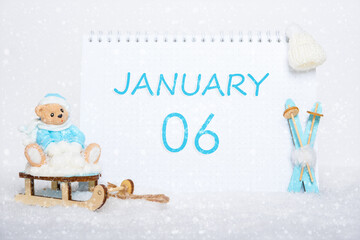 January 6th. Teddy bear sitting on a sled, blue skis and a calendar date on white snow. Day 6 of month.