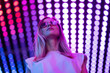 Teen hipster girl in stylish glasses standing on purple tunnel with neon light wall background, female teenager fashion model pretty young woman looking at night club city light glow