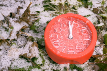 A red clock with white hands at 12 o'clock and 1 second with snowflakes on the glass lies on the green grass covered with white snow on a winter morning