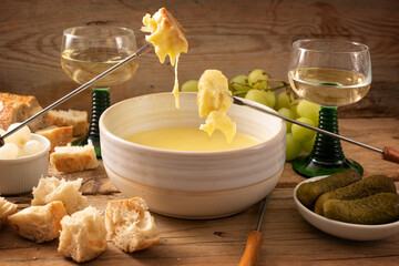 Swiss fondue from melted cheese with bread on long forks, pickles, grapes and wine on a rustic wooden table, often served at holidays like new year, selected focus