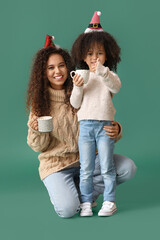 Poster - African-American woman and her little daughter with cups of hot cocoa on green background