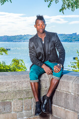 Wall Mural - Dressing in a black blazer,  green pants, leather shoes, a young black guy with mohawk hair is sitting on a rocky fence by a river, relaxing..