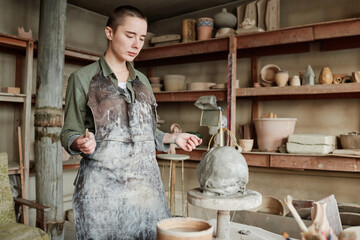 Wall Mural - Young woman in apron standing and looking at her sculpture, she learning to make it from clay in ceramic studio