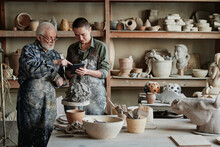 Senior Sculptor Pointing At Tablet Pc And Showing His Assistant The Ceramic Sculpture During Their Work In Pottery Studio