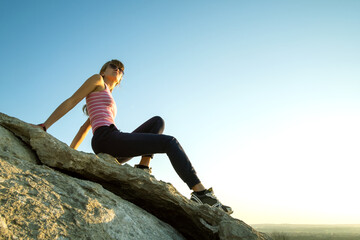 Wall Mural - Woman hiker sitting on a steep big rock enjoying warm summer day. Young female climber resting during sports activity in nature. Active recreation in nature concept.