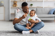 Happy Young Black Dad Spoon Feeding His Adorable Baby Son At Home
