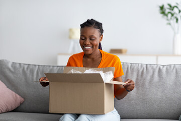 Online shopping and delivery concept. Cheerful black woman unboxing cardboard package, satisfied with purchase at home