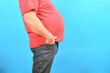 A fat man with a big belly in jeans and a red T-shirt on a blue background holds his hands in his pockets.