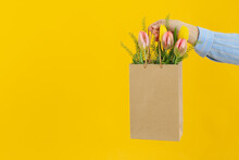 Female Hand Holds Paper Bag With Flowers On Yellow Background