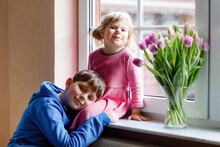 Little Girl And Kid Boy Sitting By Window With Tulip Flowers Bouquet. Happy Children, Siblings, Cute Brother And Sister, Indoors. Mother's Day, Valentine's Day Or Birthday Concept.