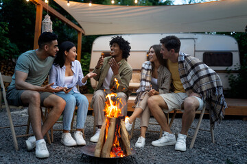 Happy multinational friends sitting near campfire, talking to each other, enjoying warm summer evening next to RV