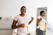 © Prostock-studio - Plus size black woman measuring waist with tape in front of mirror, showing results of slimming diet, gesturing thumb up