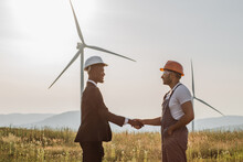 African American Man In White Helmet And Black Suit Shaking Hands With Indian Engineer With Wind Turbines On Background. Two Colleagues Making Deal About Successful Cooperation.