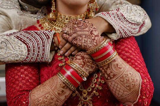 Indian bride in red wedding costume holds groom's hands on her chest while he hugs her from behind Henna arms bangles and bracelet Jewelry Hindu Bride and groom Cropped photo