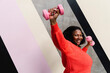 © VISTA by Westend61 - Happy plus size woman exercising with dumbbells near patterned wall