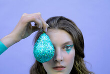 Teenage Girl With Makeup Holding Glittered Pear In Front Of Eye Against Lavender Background
