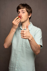 Young handsome tall slim white man with brown hair biting strawberry pointing at you in light blue shirt on grey background