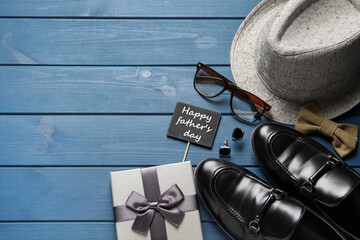 Wall Mural - Card with phrase Happy father's Day, shoes, gift box and different men accessories on blue wooden background, flat lay. Space for text