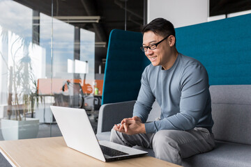 Asian with glasses, working in the office, making a video call with a laptop, smiling and rejoicing, communicating remotely with colleagues online, business man working in the office