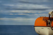 Lifeboat hanging above the decks onboard a cruise ship. Orange and white raft for use in a nautical emergency. With view of the sea with a moody sky. With space for text.