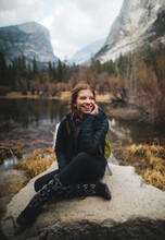 Smiling Woman In Dark Bubble Jacket Sitting On Rock Formation Near Lake