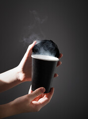 Wall Mural - a black paper cup with aroma coffee or other drink in hands on a dark background with morning light. The concept of a cheerful morning