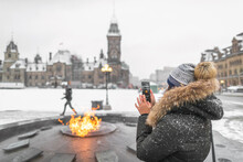 Ottawa Travel Tourist Woman Taking Photo With Phone Of Canadian Parliament In Ontario, Canada. Snowing Landscape And Centennial Flame That Do Not Freeze In Winter.