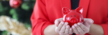 Woman In Red Jacket Holds Piggy Bank Against The Background Of Christmas Tree.