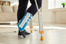 Close Up Leg Of Man Walking With Crutches In Ankle Brace With Support Adjustable Strap Fracture Fixator. Cropped Image Of Low Angle Leg Of Man Who Has Leg Injury And Walks With Crutches Room At Home