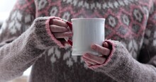 Cold Female Hands Hold White Mug Of Hot Drink With Steam Rising Under Snow Fall In Wind Outdoors. Woman Wearing Scandi Pattern Sweater And Fingers Shake From Cold And Try To Warm By Hot Cup