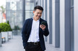 © Liubomir - asian male freelancer walking near business center holding phone, smiling reading news, successful businessman