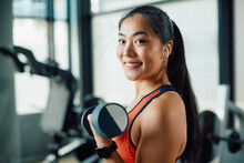Happy Asian Athletic Woman Lifts Hand Weights During Sports Training At Gym And Looks At Camera.