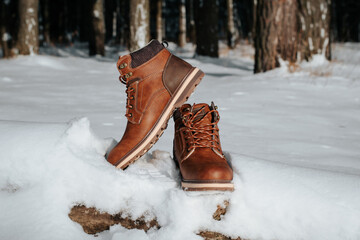 Pair of men's leather brown winter boots standing on log in snowy forest on sunny day, outdoors. Shoes for hiking and traveling. Garment object, no people