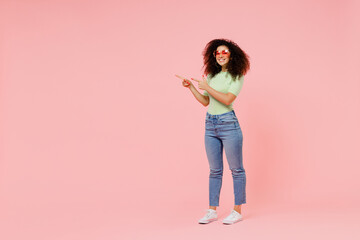 Canvas Print - Full size body length happy young curly latin woman 20s wears casual clothes sunglasses point aside on workspace area copy space mock up isolated on plain pastel light pink background studio portrait.