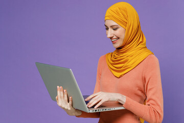 Wall Mural - Smiling fun young arabian asian muslim woman in abaya hijab yellow clothes hold use work on laptop pc computer type browsing chatting isolated on plain pastel light violet background studio portrait.