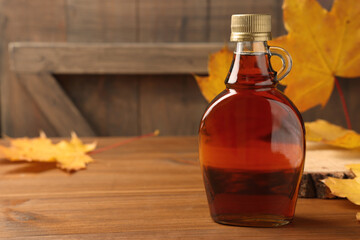 bottle of tasty maple syrup and dry leaves on wooden table, space for text