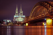 The Illuminated Cologne Cathedral On The Rhine And The Hohenzollern Bridge In Cologne At Night With Purple Sky.