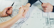 © Andrey - Two engineers work together and use a tablet to search for blueprints and analyze an architectural plan on a table. Agreement by agreement by shaking hands.