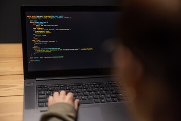 Over shoulder view of woman using modern laptop for typing data code while sitting at desk during evening time at home. Focus on screen with web programs.
